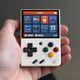 The Miyoo Mini, showing its home screen on the display, being held in someone's left hand.